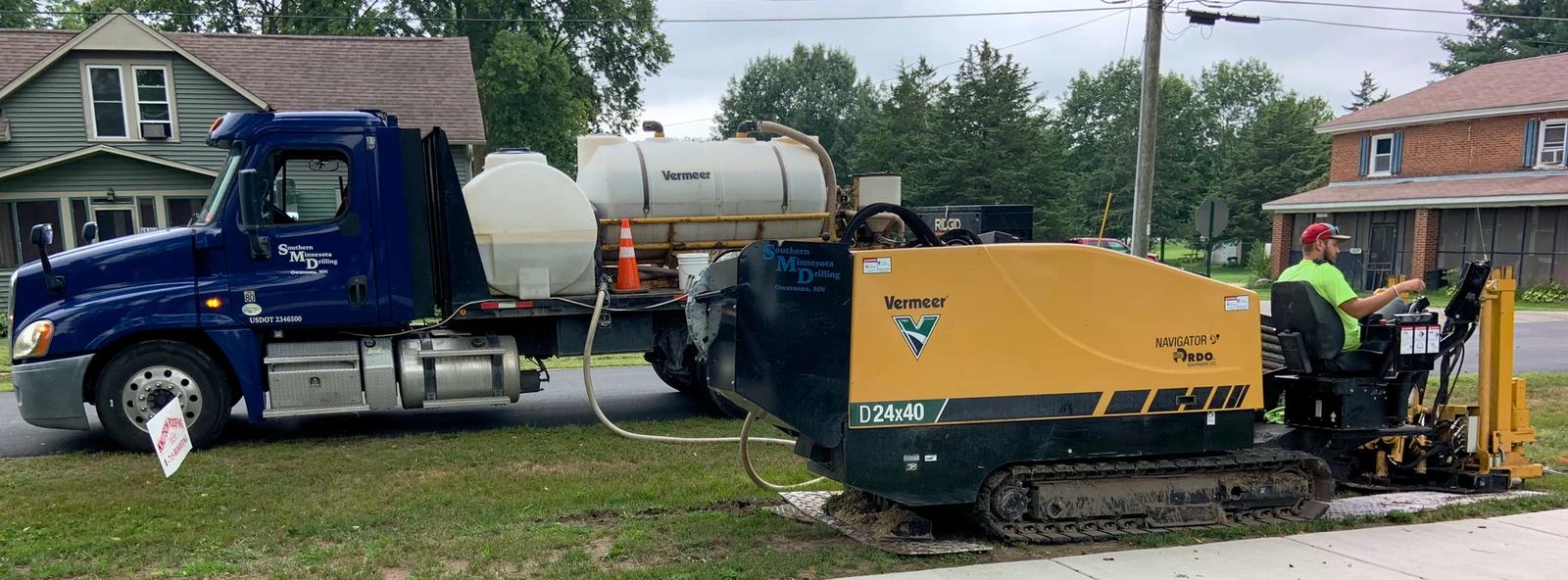 Een gele machine voor gestuurde boringen op een grasstrook naast een straat, met een vrachtwagen erachter