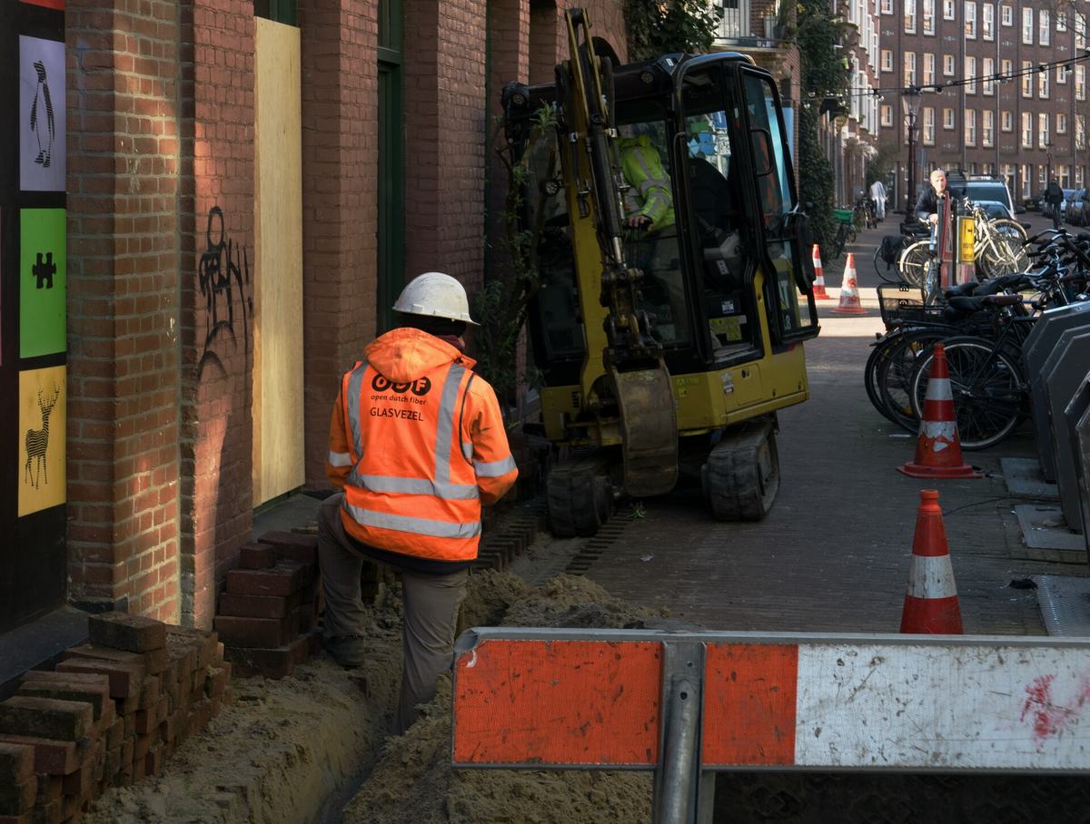Een wegenwerker in een oranje jas bij een minigraver, in een opengebroken voetpad in een stadsstraat