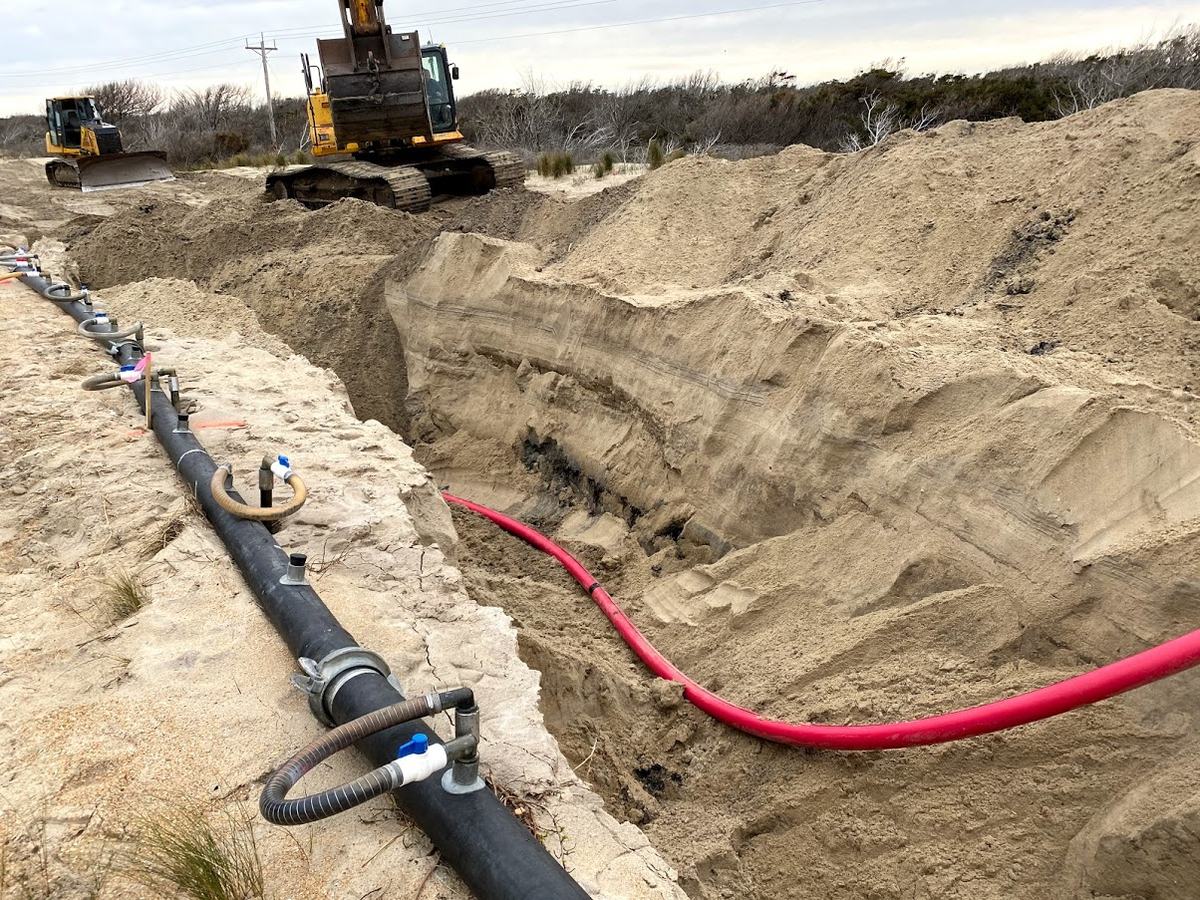 Een open sleuf in het zand met een zwarte buis en een rode kabel, een graafmachine op de achtergrond